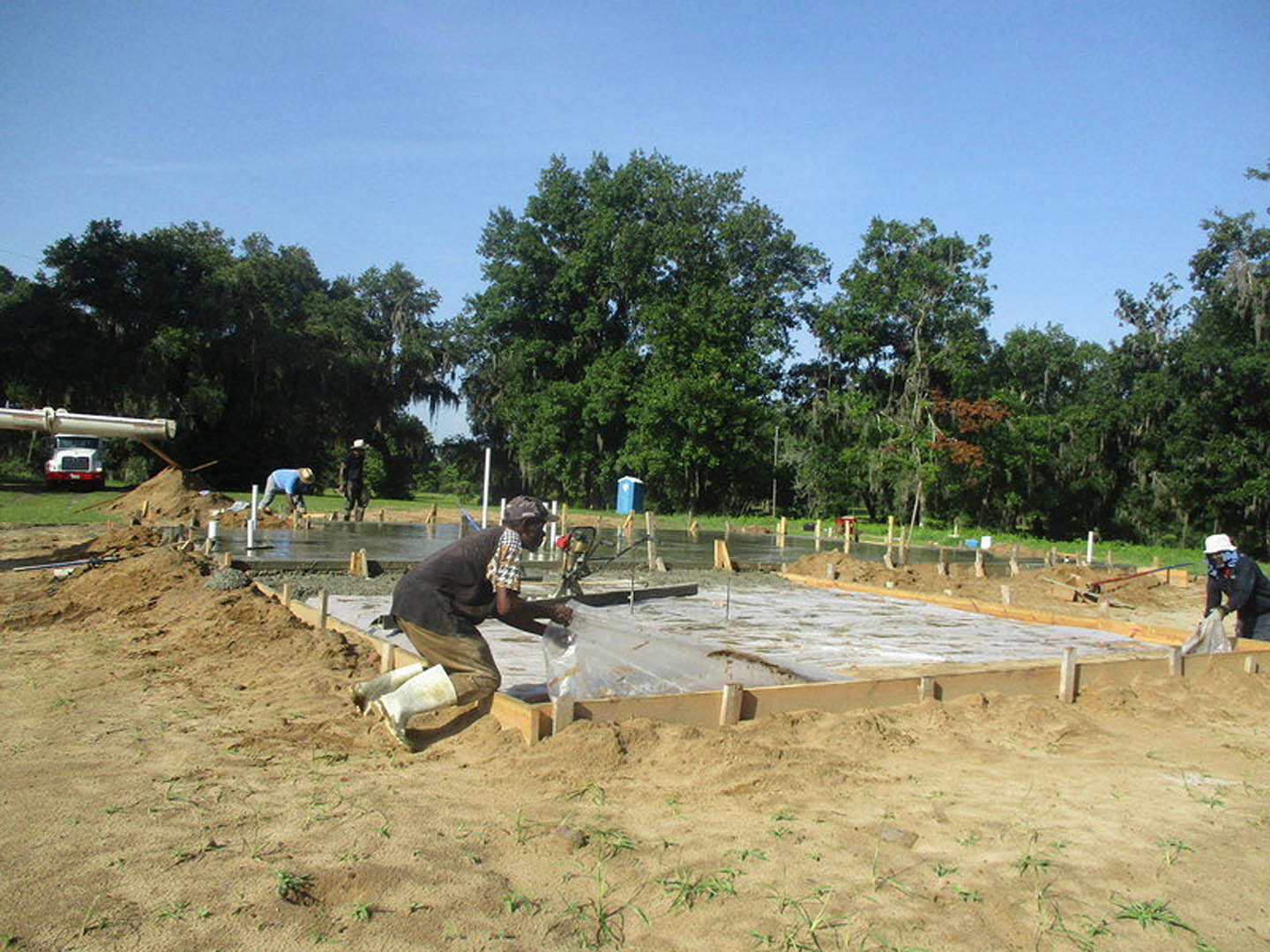 Man in hat and boots pouring concrete into a foundation on muddy construction site, truck parked nearby, grassy field and trees in background