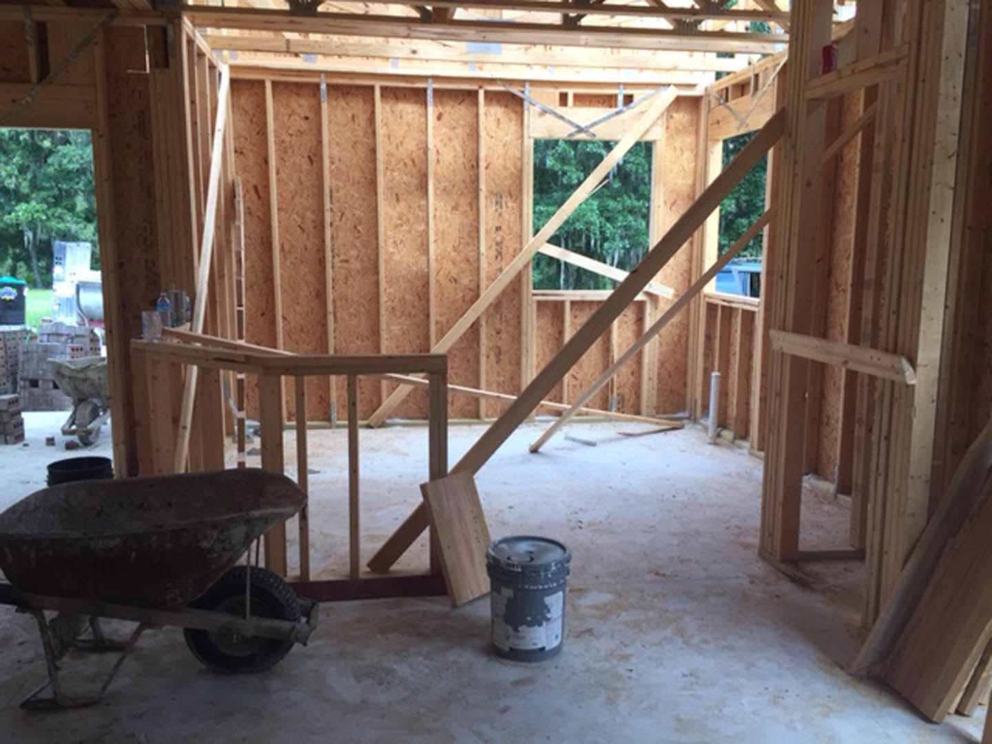 Unfinished room with exposed wooden beams, concrete floor, wheelbarrow, and paint bucket, showing construction materials and remodeling progress