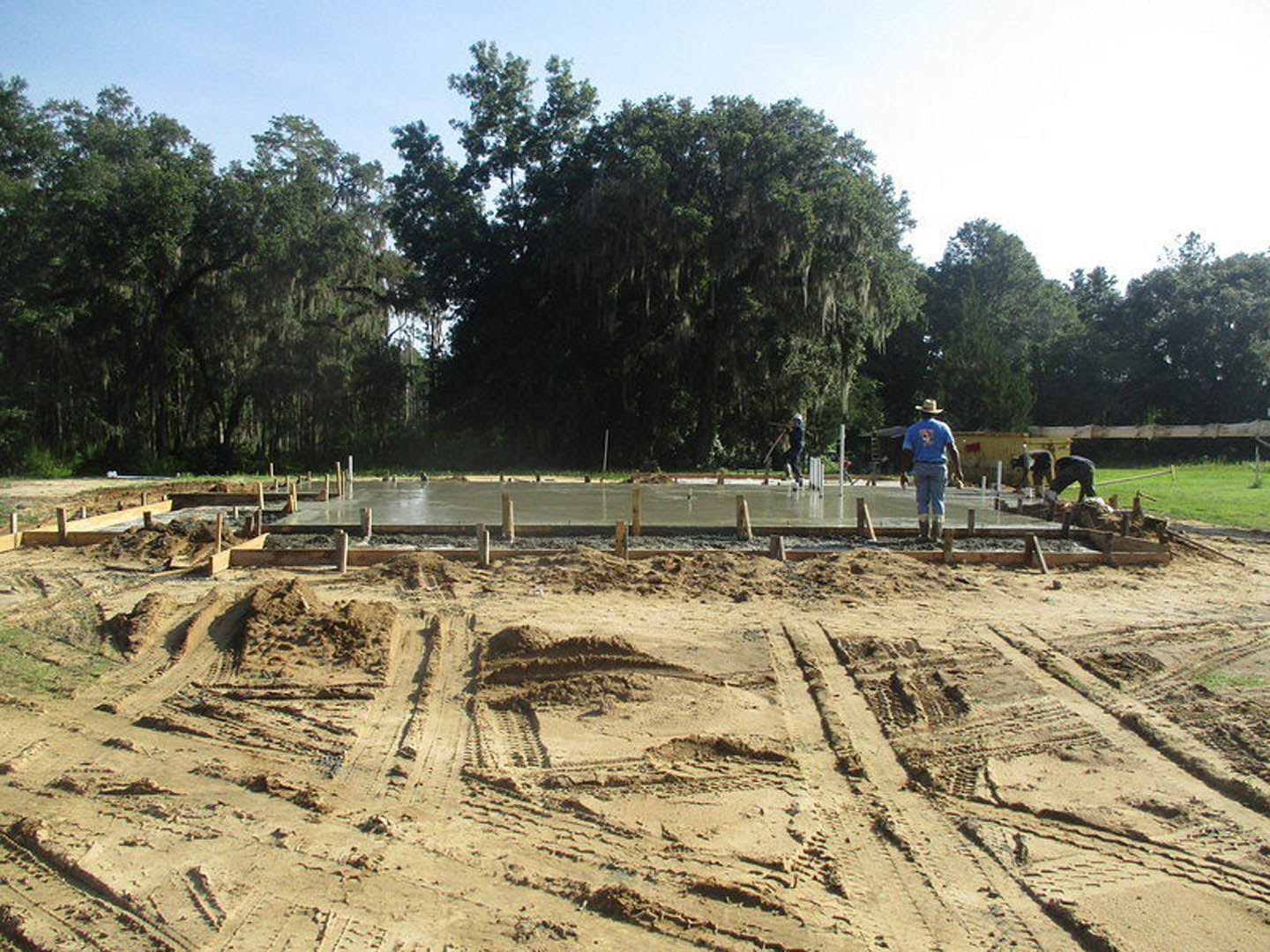 Concrete foundation under construction with workers in blue shirts, sandy soil with tire tracks, grassy area being mowed, moss-covered tree in background