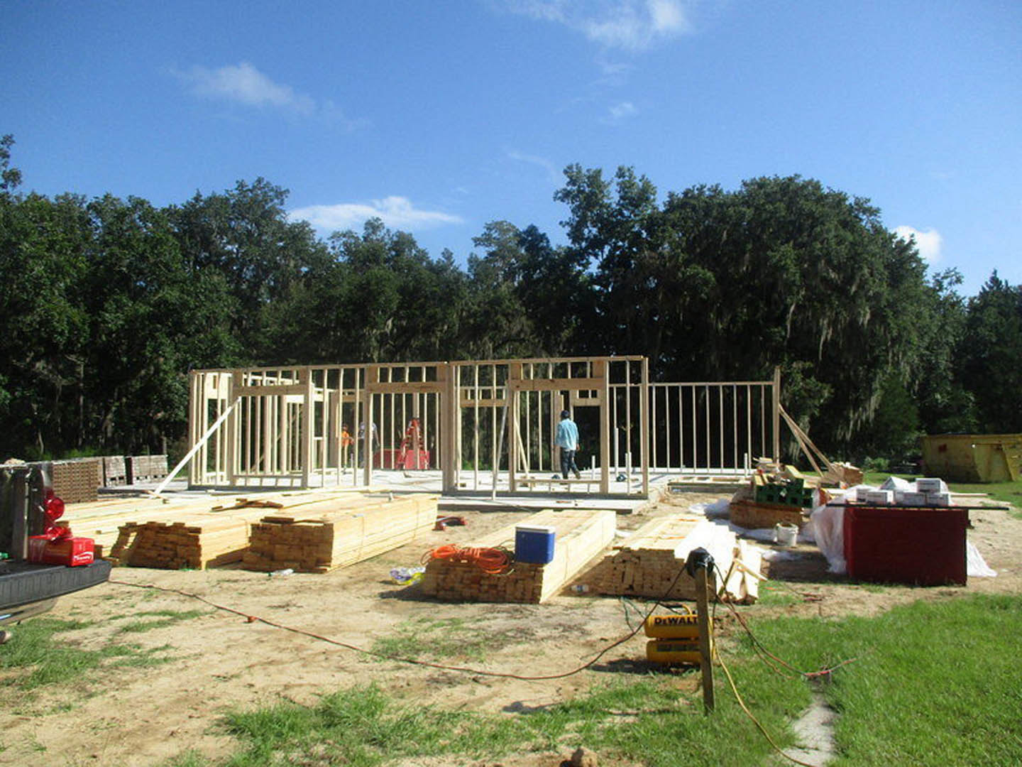 Partially built house with exposed framing, red wall section, white fence in foreground, blue cooler on grass, two men working near construction materials, trees and cloudy sky in