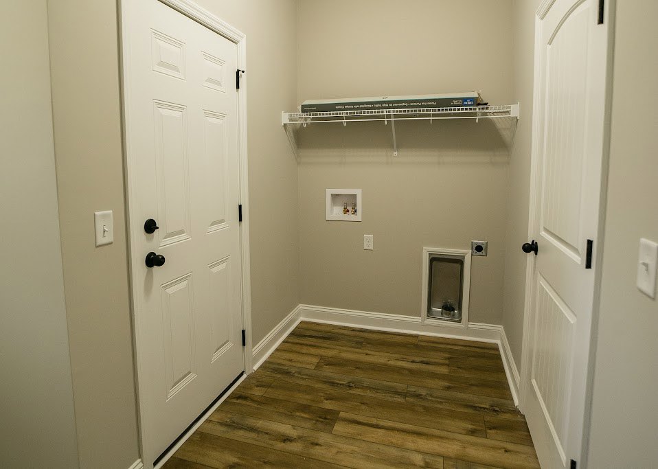 Wood plank flooring, white baseboards, white door with black knobs, white shelving with small bottles, plaster walls and ceiling