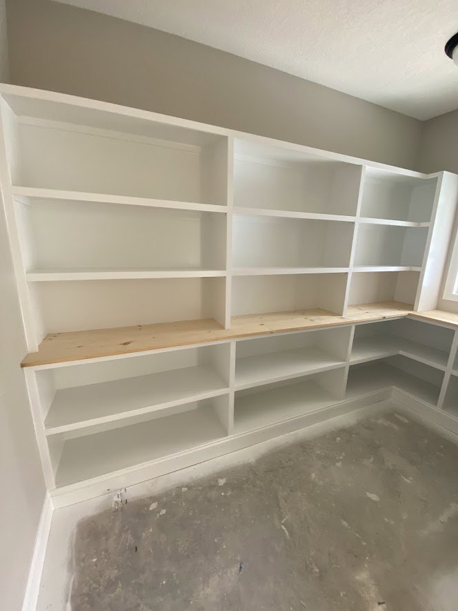 White built-in shelving with wooden top and black accent line against plaster wall, concrete floor, natural light streaming in
