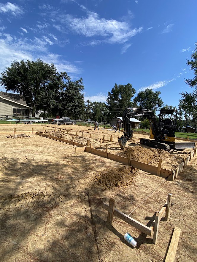 Bulldozer parked beside a partially built house with wooden framing, surrounded by dirt ground, temporary fencing, and trees under a blue sky with scattered clouds