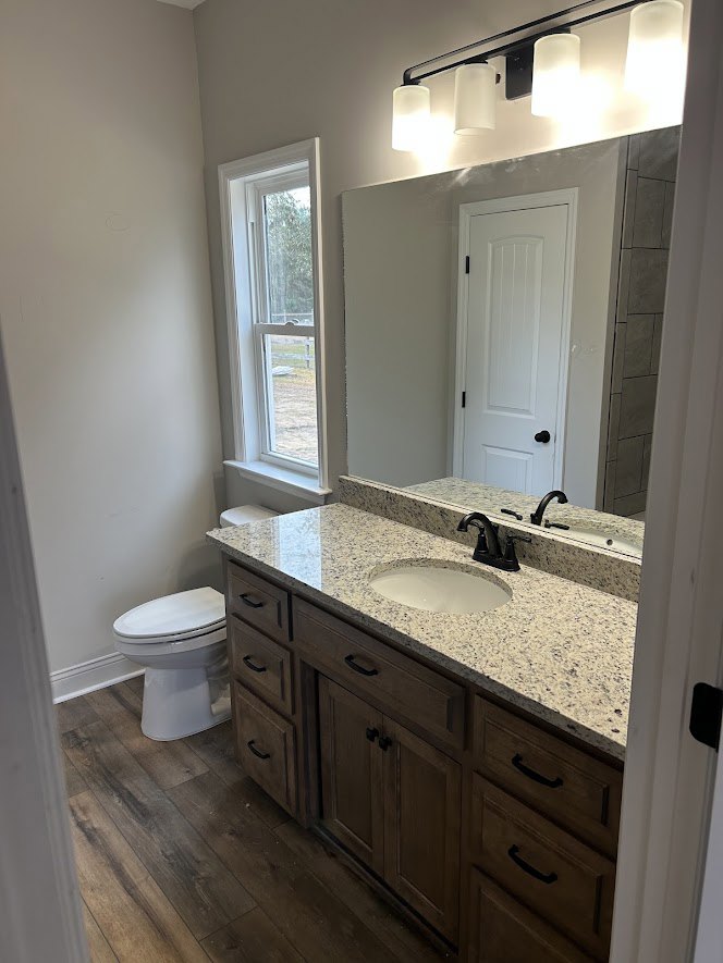 Bathroom with marble countertop, white sink and cabinet, closed white toilet, wood flooring, and wall-mounted mirror