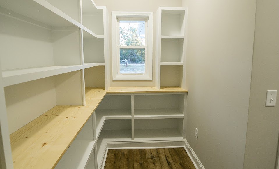 Built-in white shelves against a plaster wall, wood floor with white trim, large window overlooking fenced yard and trees