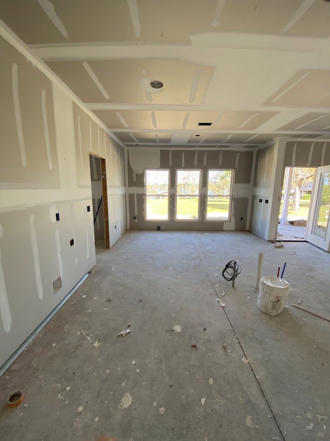 White-walled room with multiple windows, bucket of paint and plastic bag on unfinished floor, view of grassy field and neighboring house through windows