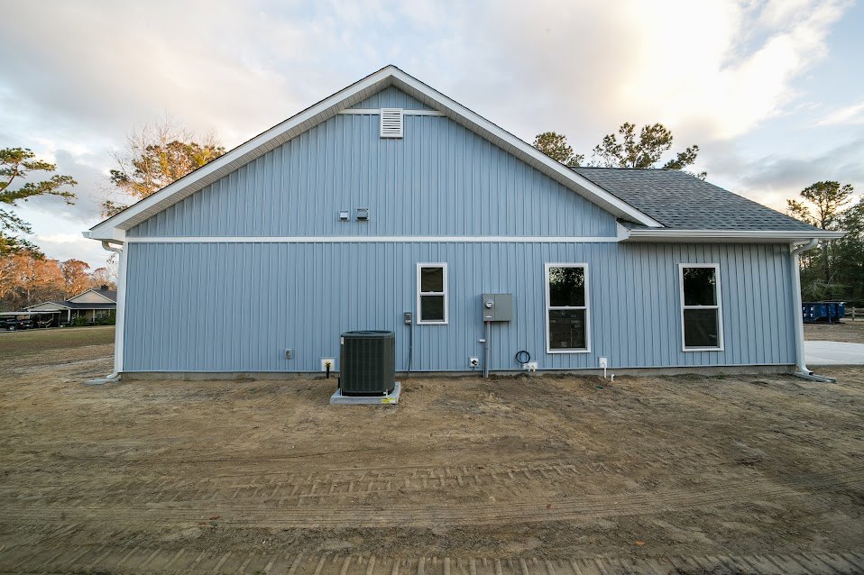 Single-story home with blue siding, white-framed windows, covered porch, dirt yard with tire tracks, black metal grill near entrance, and cloudy sky overhead.