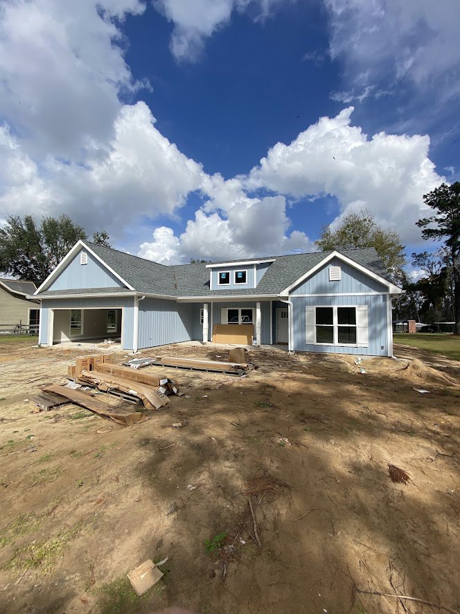 White house under construction with blue roof, white-framed window, dirt yard scattered with debris, blue sky and clouds overhead