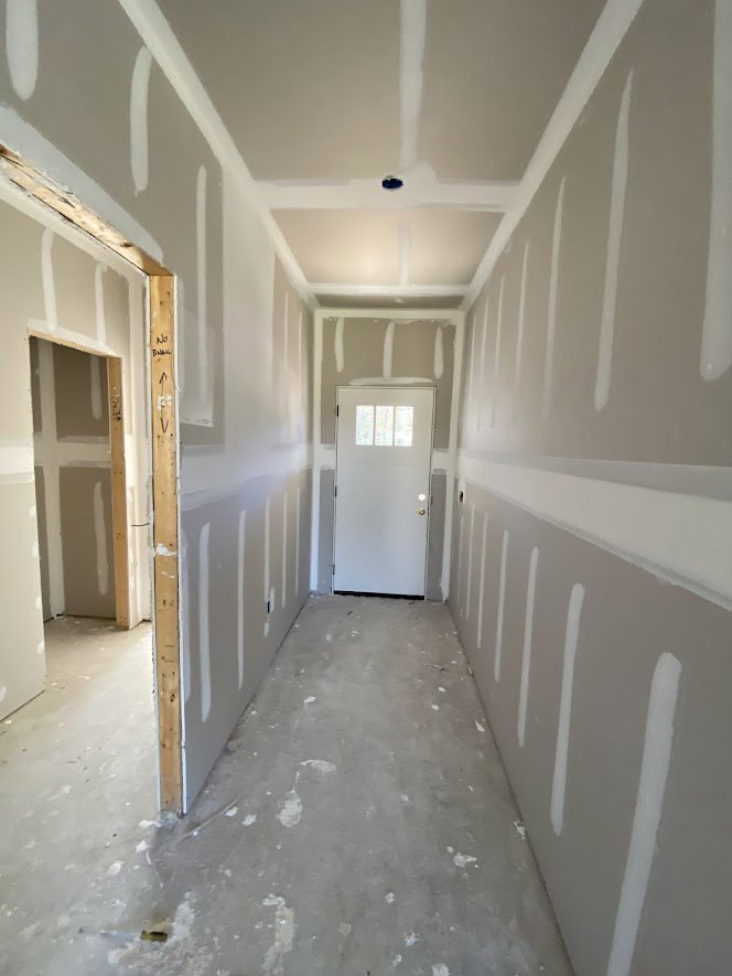 Hallway with white painted walls, white door featuring a window, wooden door frame, light-colored flooring, and ceiling fixtures