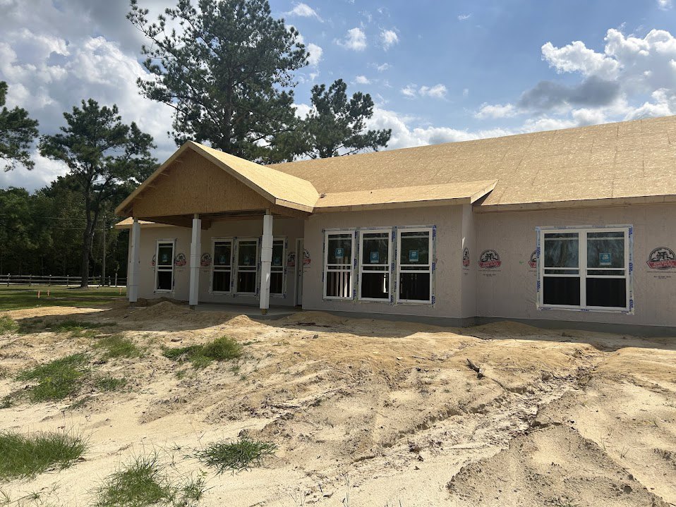 Partially built home with exposed framing, multiple windows displaying blue permit signs, sandy ground and dirt pile in foreground, mature trees and cloudy sky in background