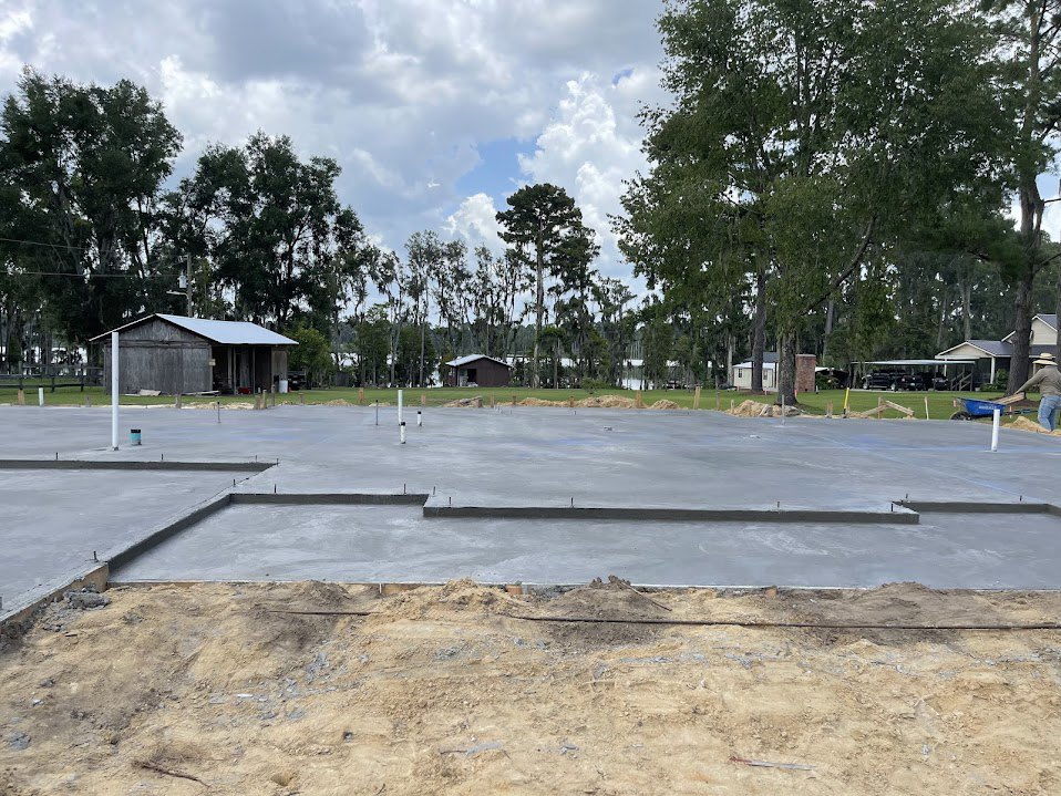 Dirt construction site bordered by mature trees, neighboring houses, a barn with metal roof, person in hat standing near large tree, overcast sky above