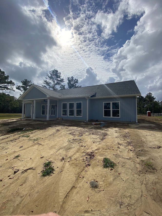 Partially built house with exposed framing, white-trimmed windows, dirt field in foreground, scattered grass and plants, cloudy sky overhead, trees in background