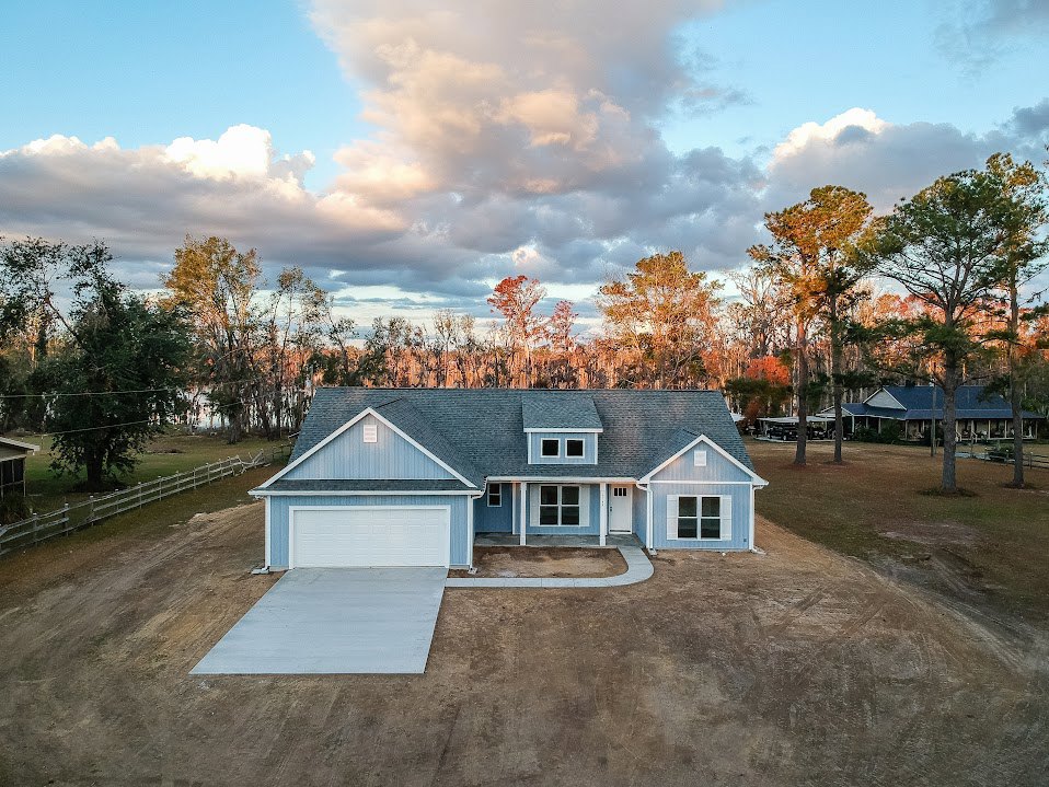Two-story house with white siding and blue trim, attached garage with white door, concrete driveway, mature trees in background, partly cloudy sky