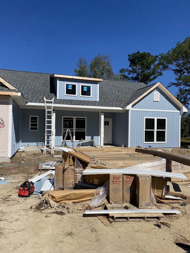 Partially built house with exposed siding, white-framed windows, blue roof, ladder leaning against exterior wall, and construction materials including boxes and a red and black bag