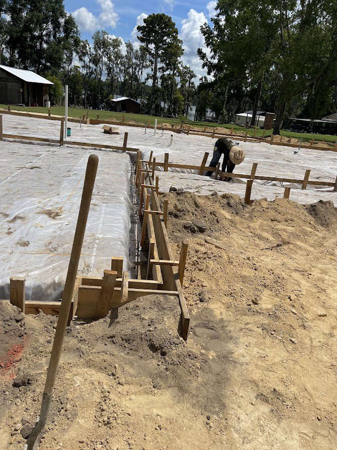 Framed house under construction with exposed roof trusses, worker in hat standing on dirt near wooden pole, mature trees and finished homes in background, cloudy sky overhead