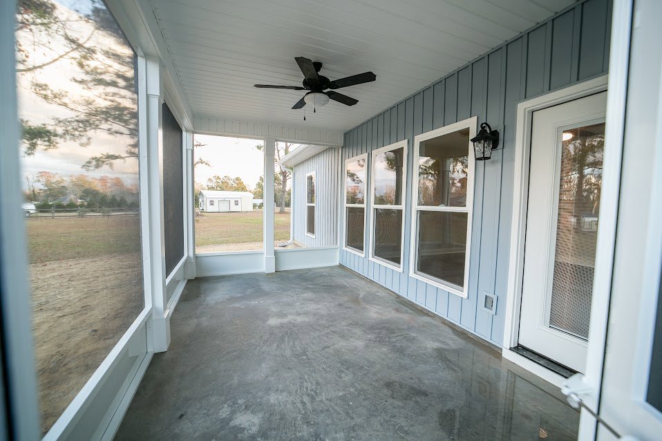 Covered porch with concrete floor, ceiling fan with light, white-framed windows with blinds, and a single door leading inside