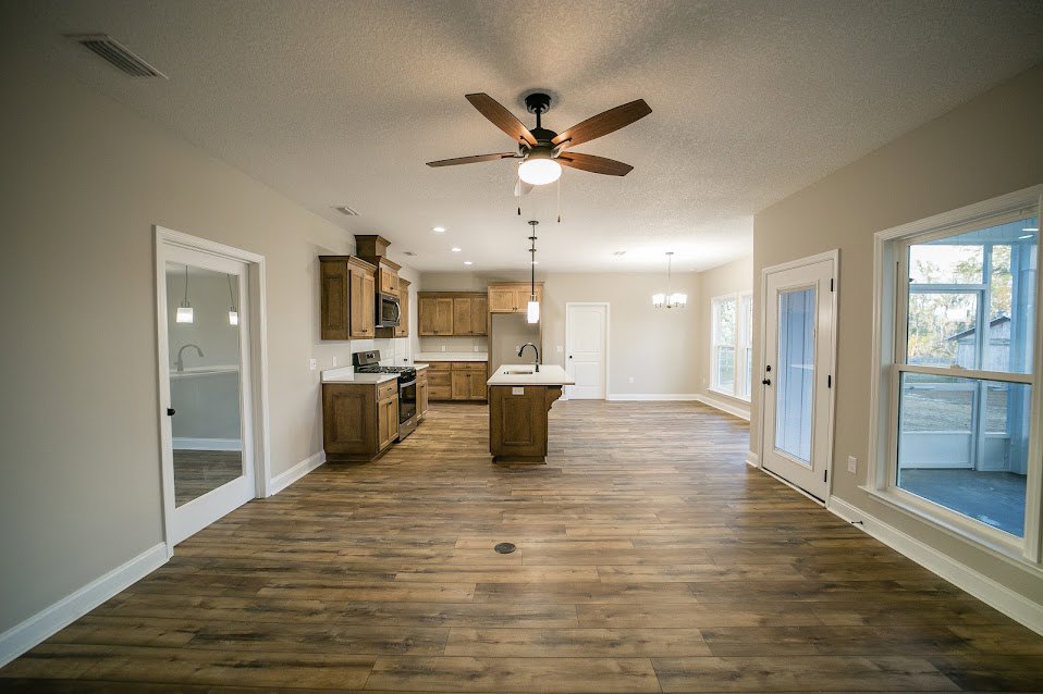 Open kitchen and dining area featuring wood flooring, central island with sink, stainless steel stove and oven, white-framed window, and ceiling fan with light fixture.