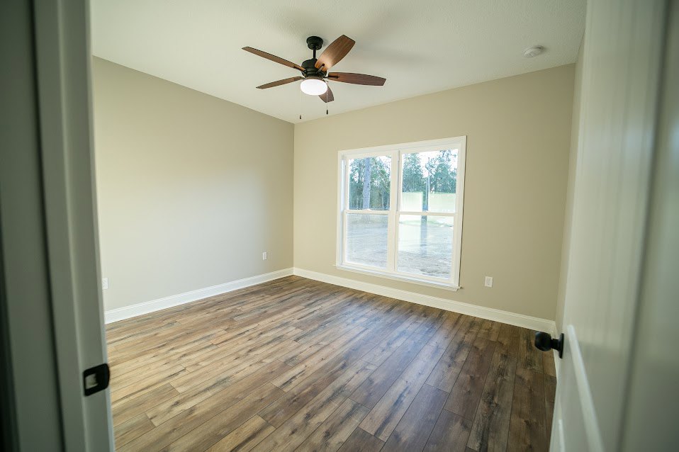 Ceiling fan with light fixture mounted on white ceiling, large window overlooking trees, wood flooring, white walls, white door