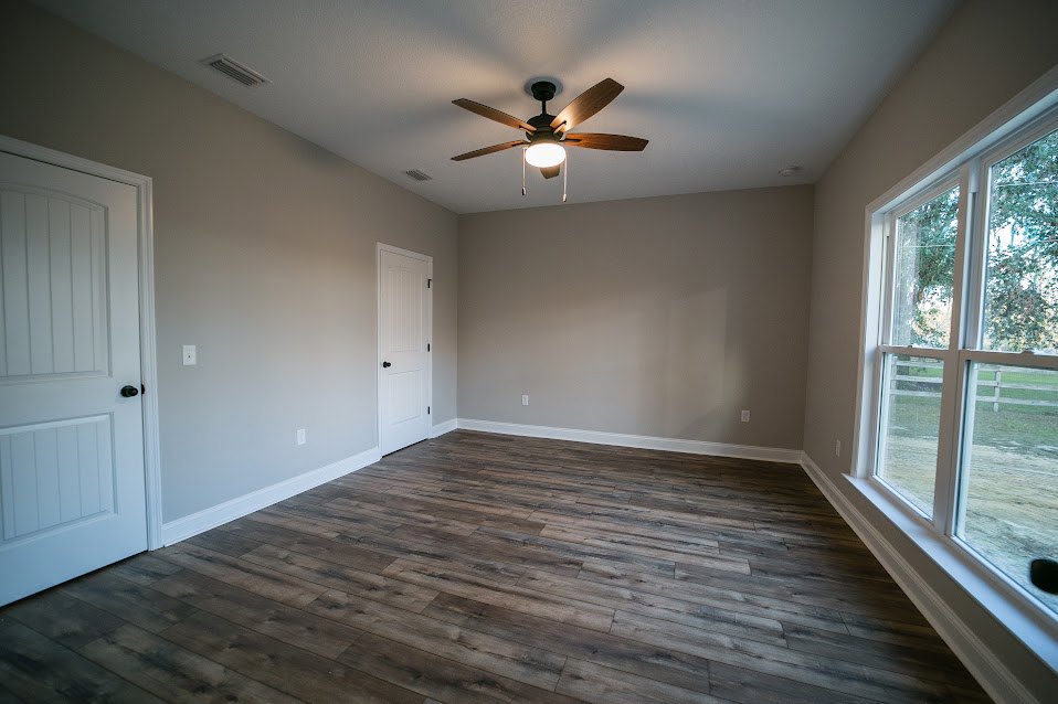 Ceiling fan with light fixture, wood plank flooring, white door with black hardware, window overlooking fenced yard, neutral painted walls