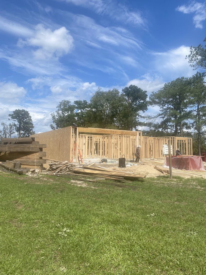 Wood-framed house under construction with exposed beams, lumber piles on grass, plastic-covered work table, and trees in the background beneath a partly cloudy sky