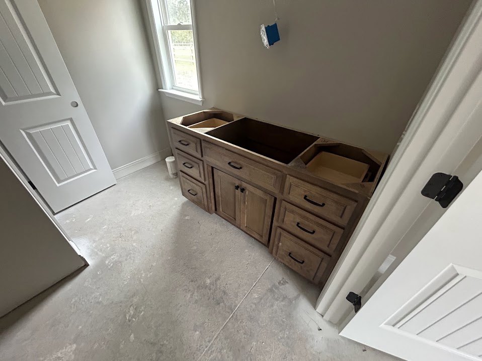 Bathroom with wood cabinet and drawers, white door featuring a hole and mounted black camera, window with white frame, light-colored flooring and walls.