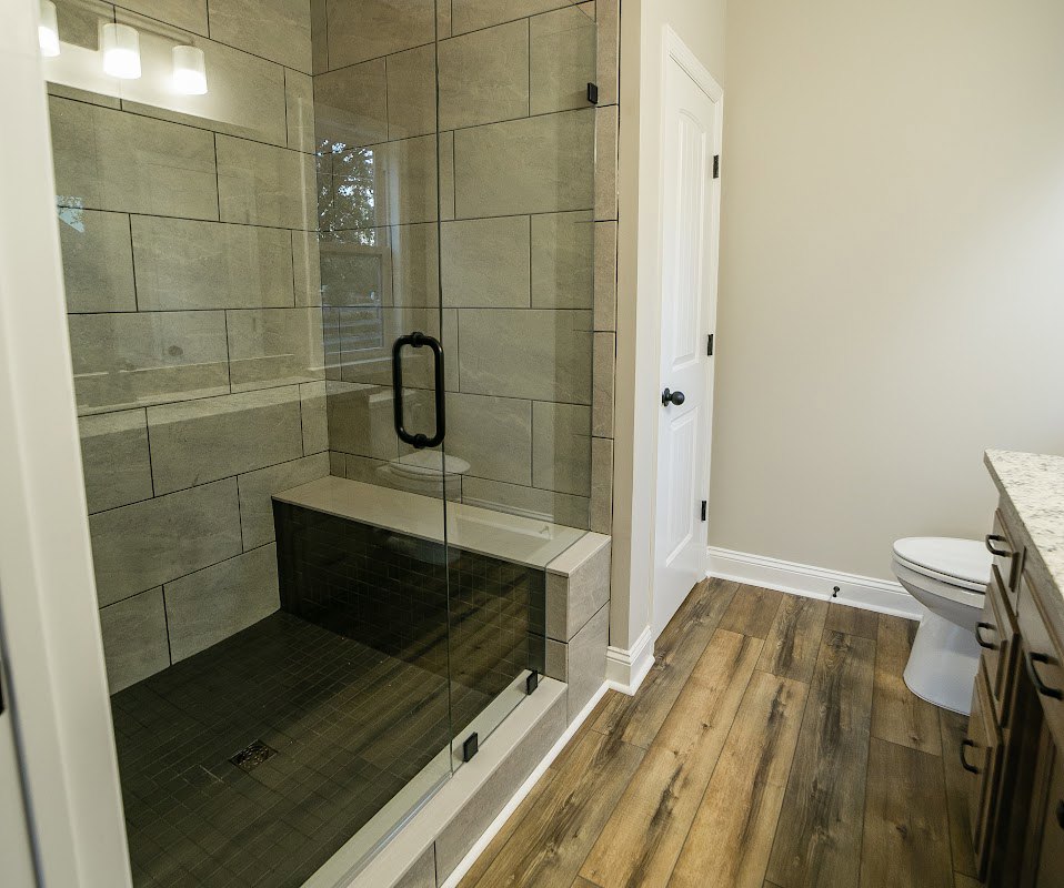 Bathroom with glass shower enclosure, white toilet, marble tile walls, wood floor, white door with black handle, and wall-mounted sink with mirror.