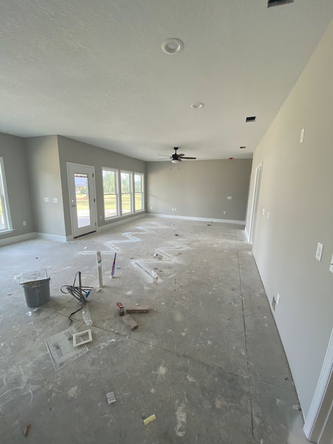 Unfinished room with plaster walls, ceiling fan, row of windows, door with glass insert, scattered tools, white frame on newspaper, and lidded bucket on floor
