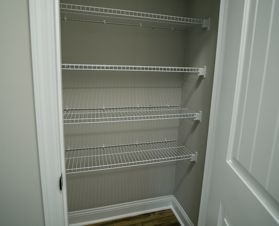 White wire shelving mounted on a closet wall above light wood flooring, with white painted walls and a partially visible door.