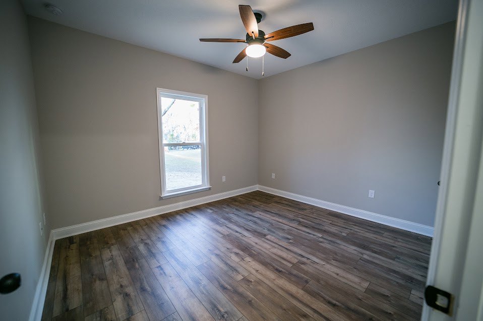 Wood-floored room with white walls, ceiling fan with light fixture, large window overlooking green yard, black and white light switch on wall