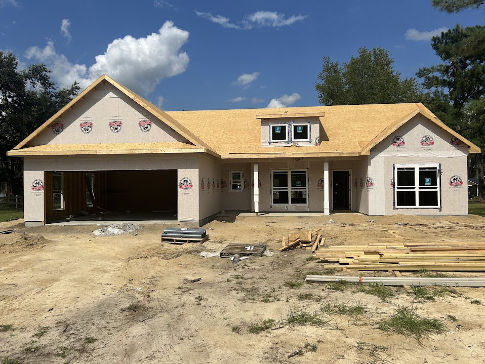 Partially built house with exposed framing, white window frame, dirt yard scattered with pallets and wood planks, blue sky with clouds overhead