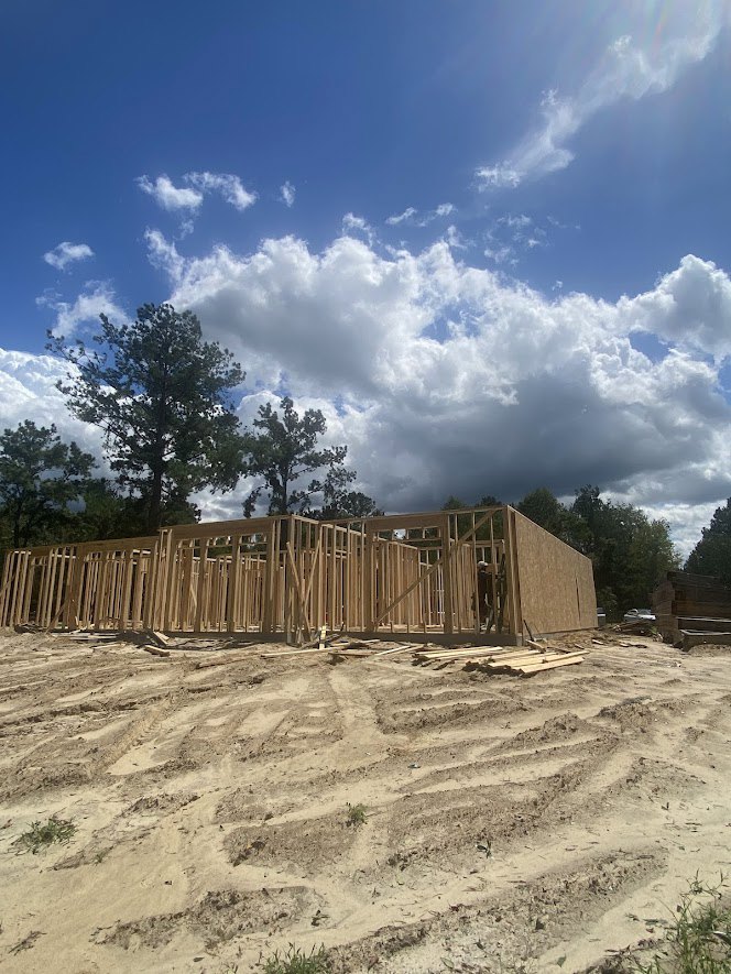 Wood-framed house under construction on dirt lot, surrounded by trees under a partly cloudy blue sky
