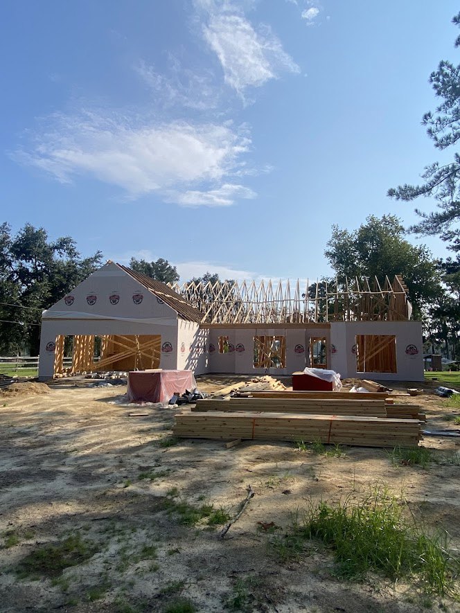 Wood-framed house under construction with exposed beams, unfinished roof, grassy lot, and surrounding trees under a cloudy sky