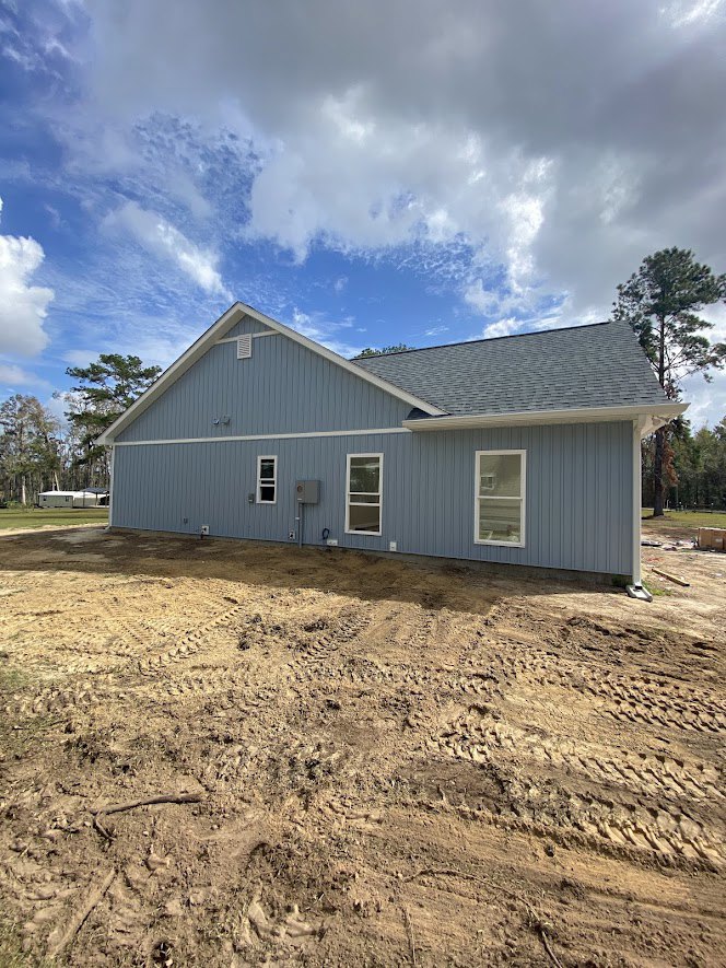 Two-story custom home with white-framed windows, gray roof, and unfinished dirt yard bordered by mature trees under a clear blue sky