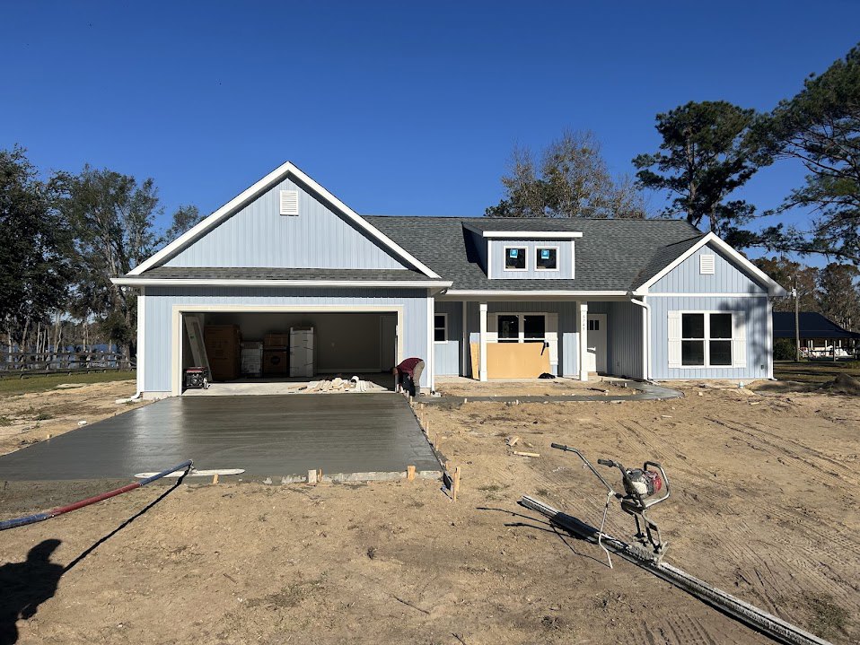 Framed custom home with attached garage, white siding, multiple windows, concrete slab foundation, construction tools and machinery on dirt lot, tree and blue sky in background