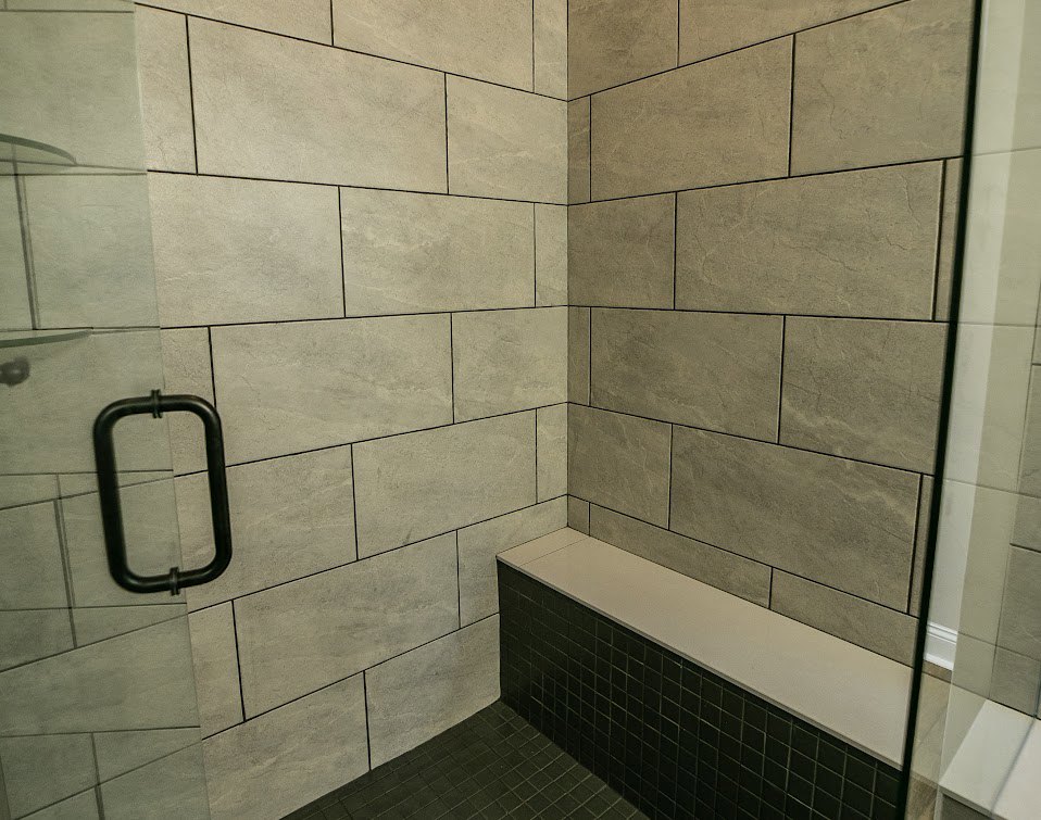 Shower with black metal-framed glass door, white countertop adjacent to tiled wall, built-in bench, mix of white and stone tile finishes