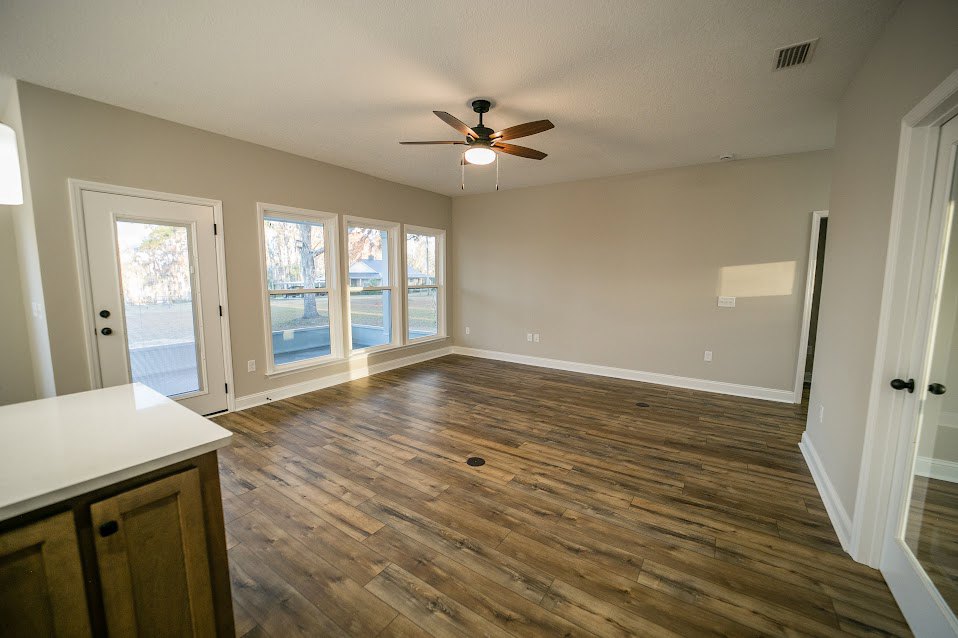 Open living area with hardwood floors, ceiling fan with light, white walls, row of windows, white door with glass insert, wood cabinetry topped with white countertop