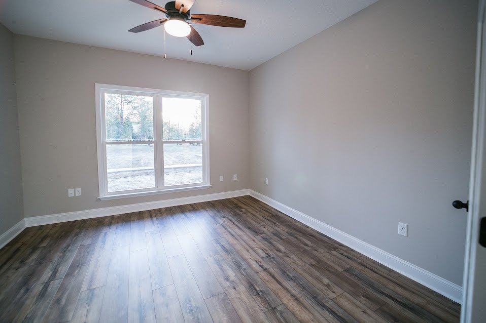 Wood floor room with white plaster walls, large window showing outdoor greenery, ceiling fan with illuminated light fixture