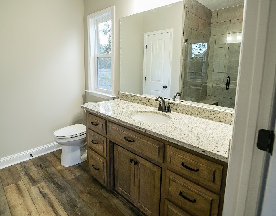 Bathroom with marble countertop, large wall mirror, white toilet, under-sink cabinets, white door with black hinges, window with white frame, glass shower enclosure with black
