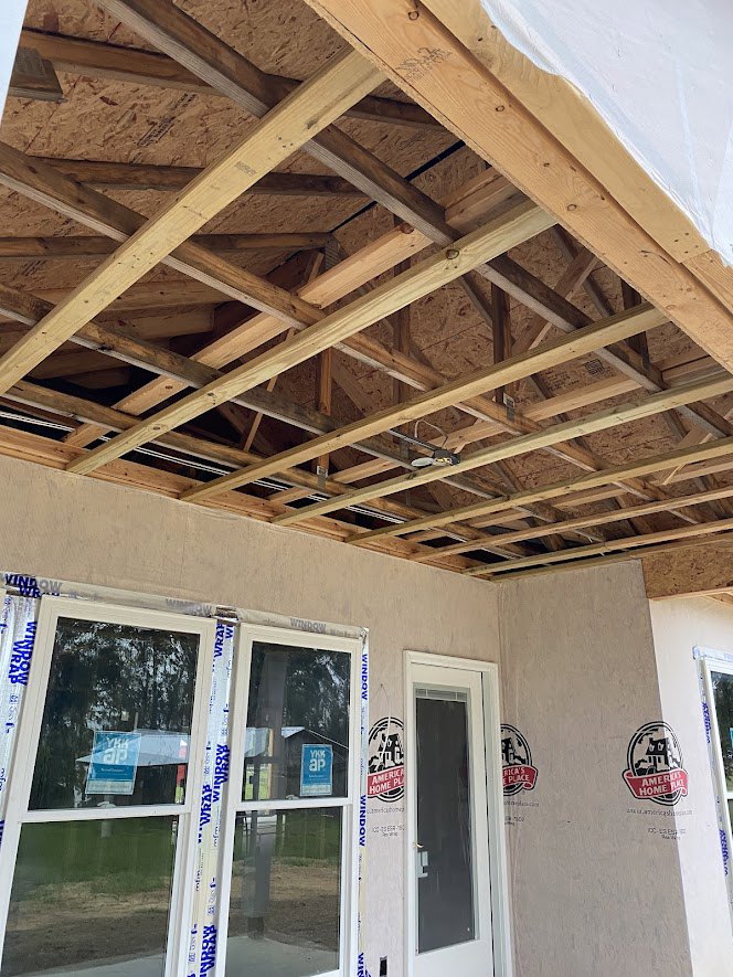 Partially built house with exposed wooden framing, white roof panels, taped window with construction sign, and visible logo on exterior surface