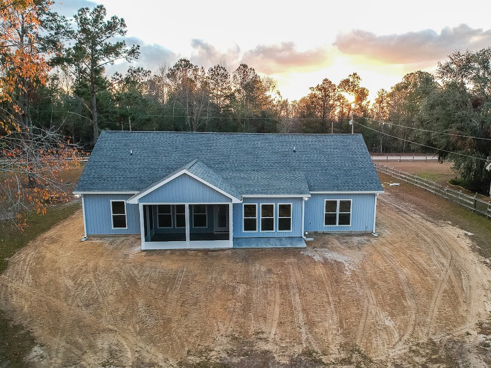 Blue house with white pillars and porch, row of windows on blue siding, dirt construction area in grassy field, wooden fence in background, cloudy sky overhead
