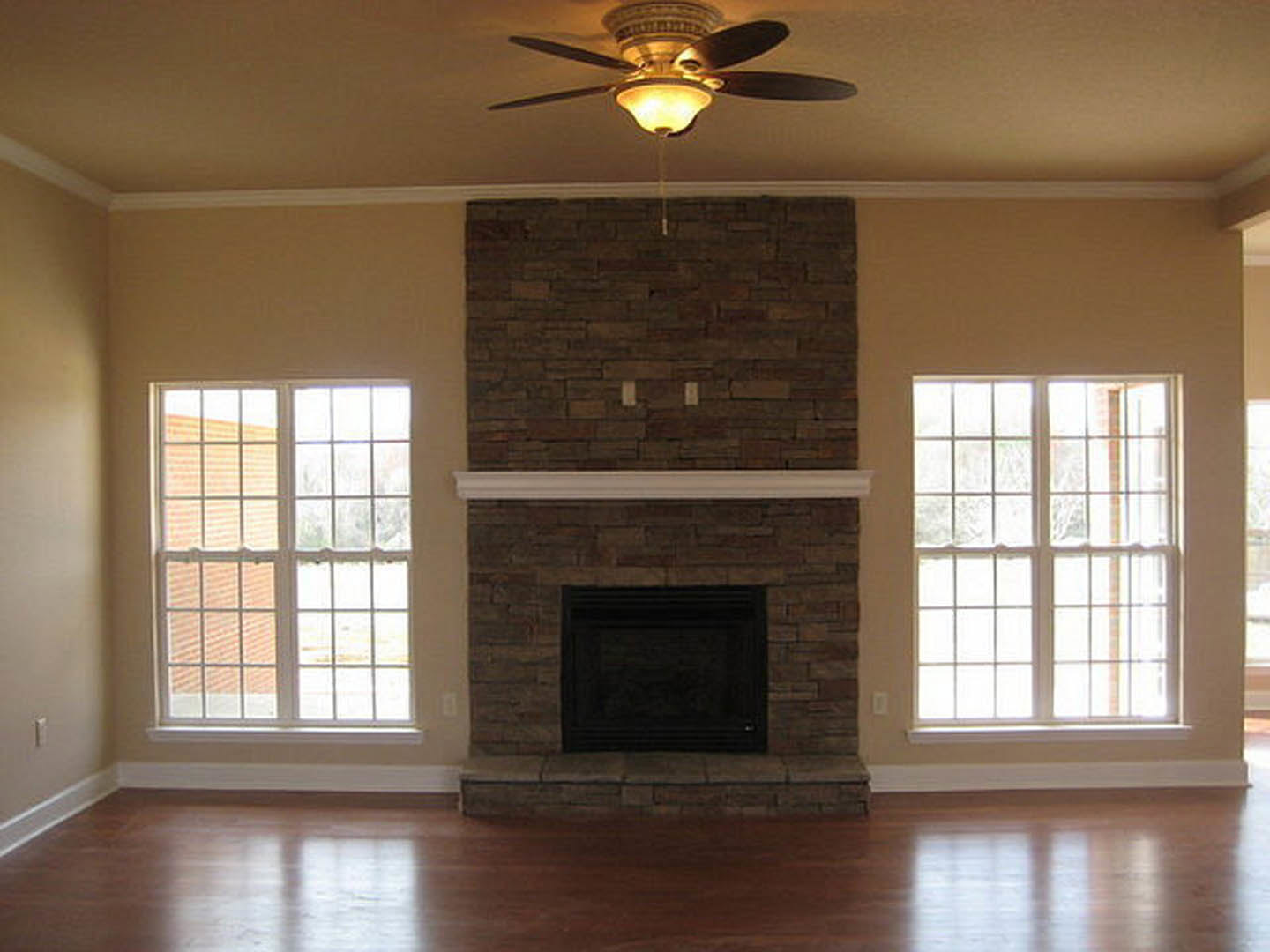 Living room featuring a white-framed window, stone fireplace with white mantel, wood flooring, ceiling fan with light, and neutral wall molding.
