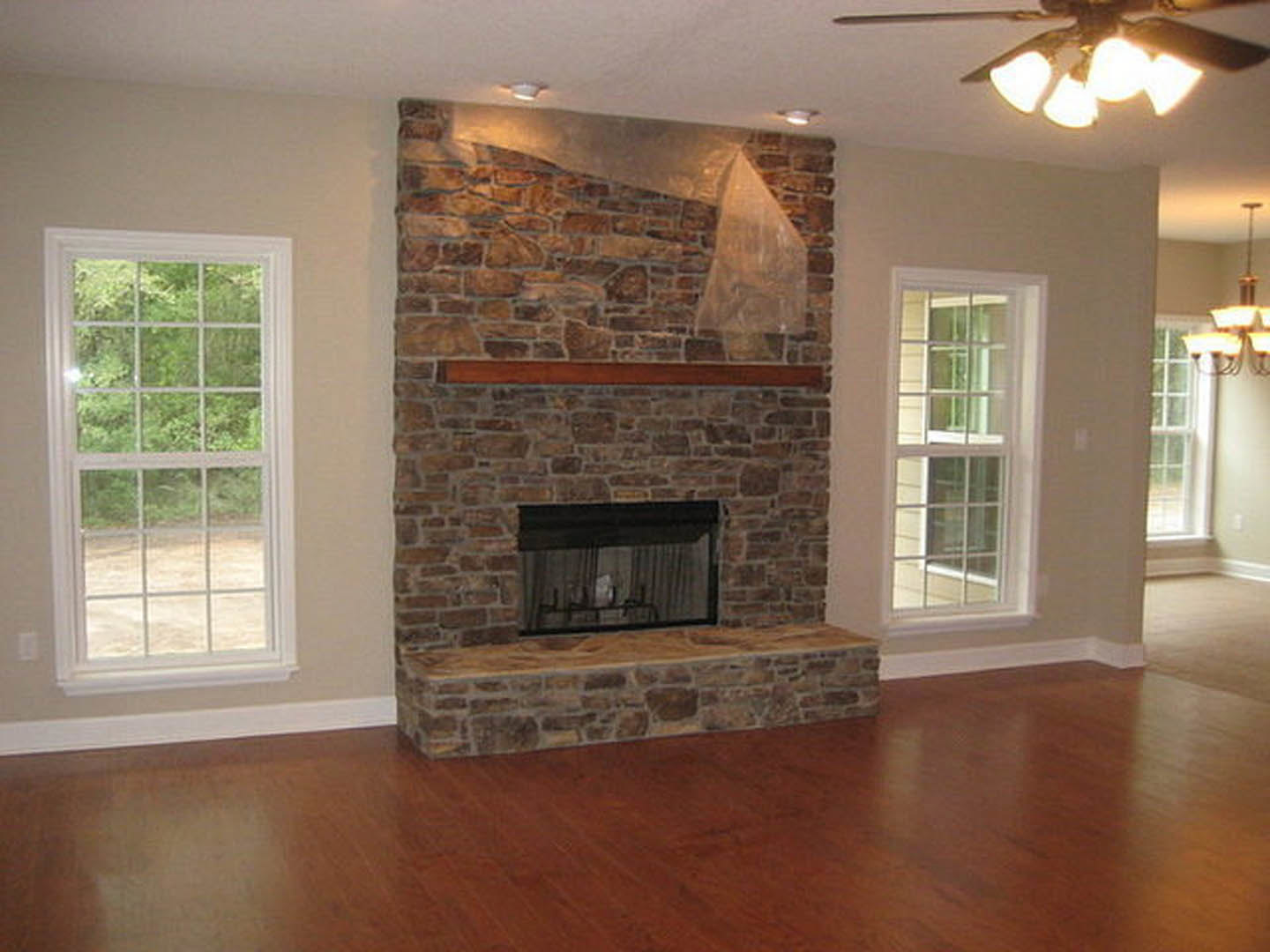 Stone fireplace with wood mantel in living room, multi-pane window with sheer curtain, hardwood floors, neutral walls, and cozy seating area.