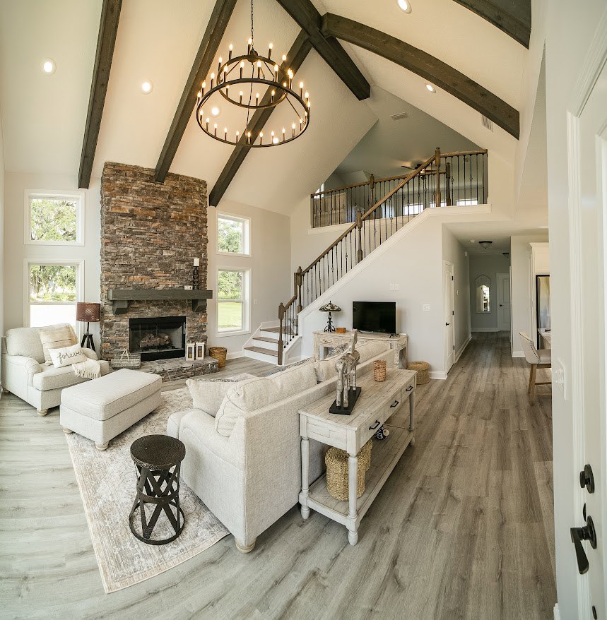 Living room with stone fireplace, multi-arm chandelier, white couch draped with blanket and pillow, metal stool, wood table with shelf and basket, person standing near large