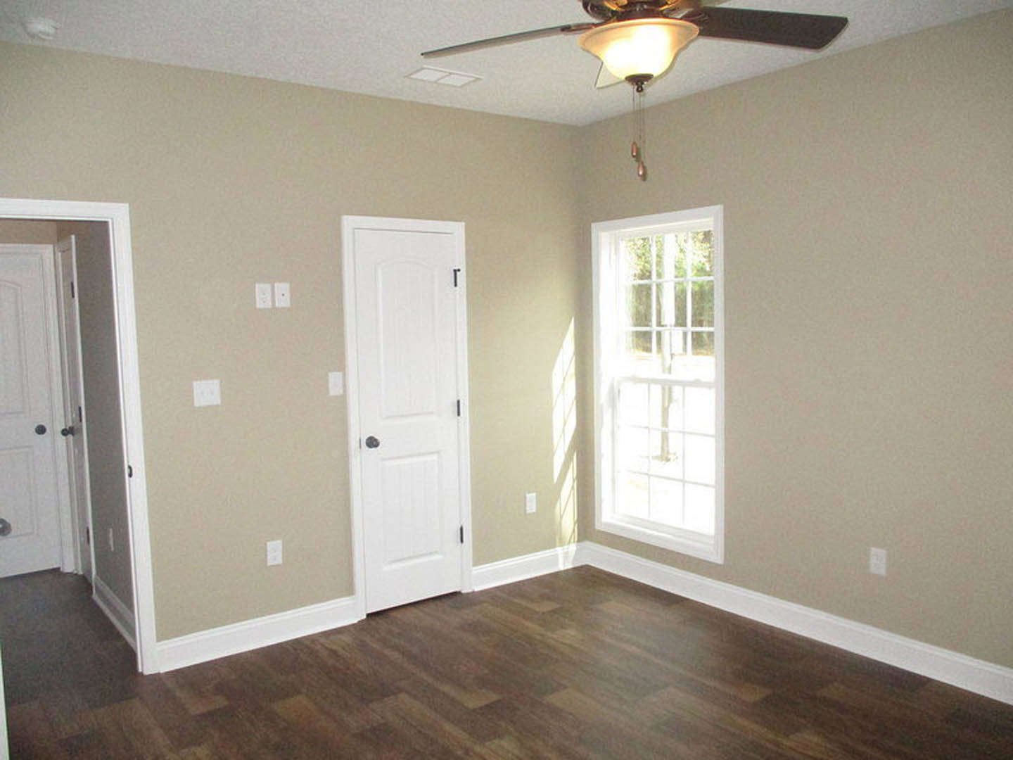 Wood floor with white trim, white door with black handle, white-framed window, ceiling fan with dark blades, smooth white walls, square wall outlet
