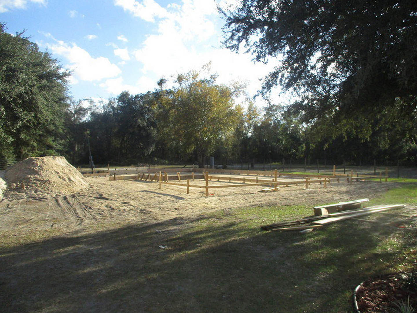 Dirt construction site with a large pile of lumber, sand mound, wooden fencing, and grassy patches under a cloudy sky
