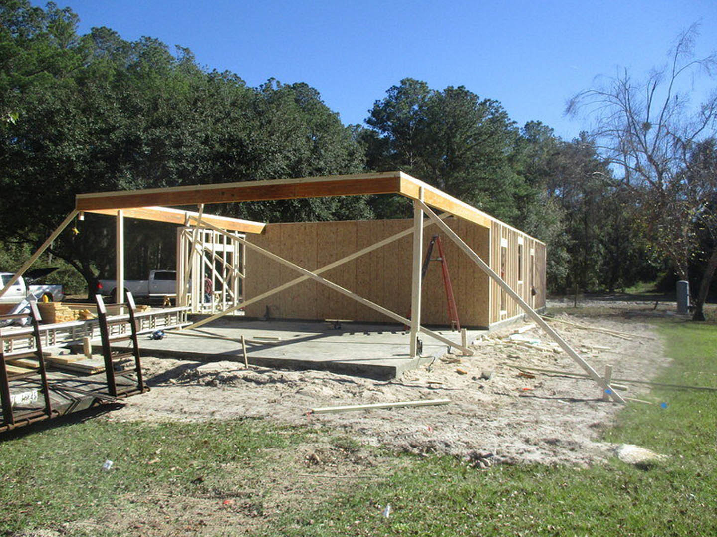 Wood-framed house under construction with exposed beams, ladder leaning against structure, leafless tree nearby, blue sky overhead, white car parked in background