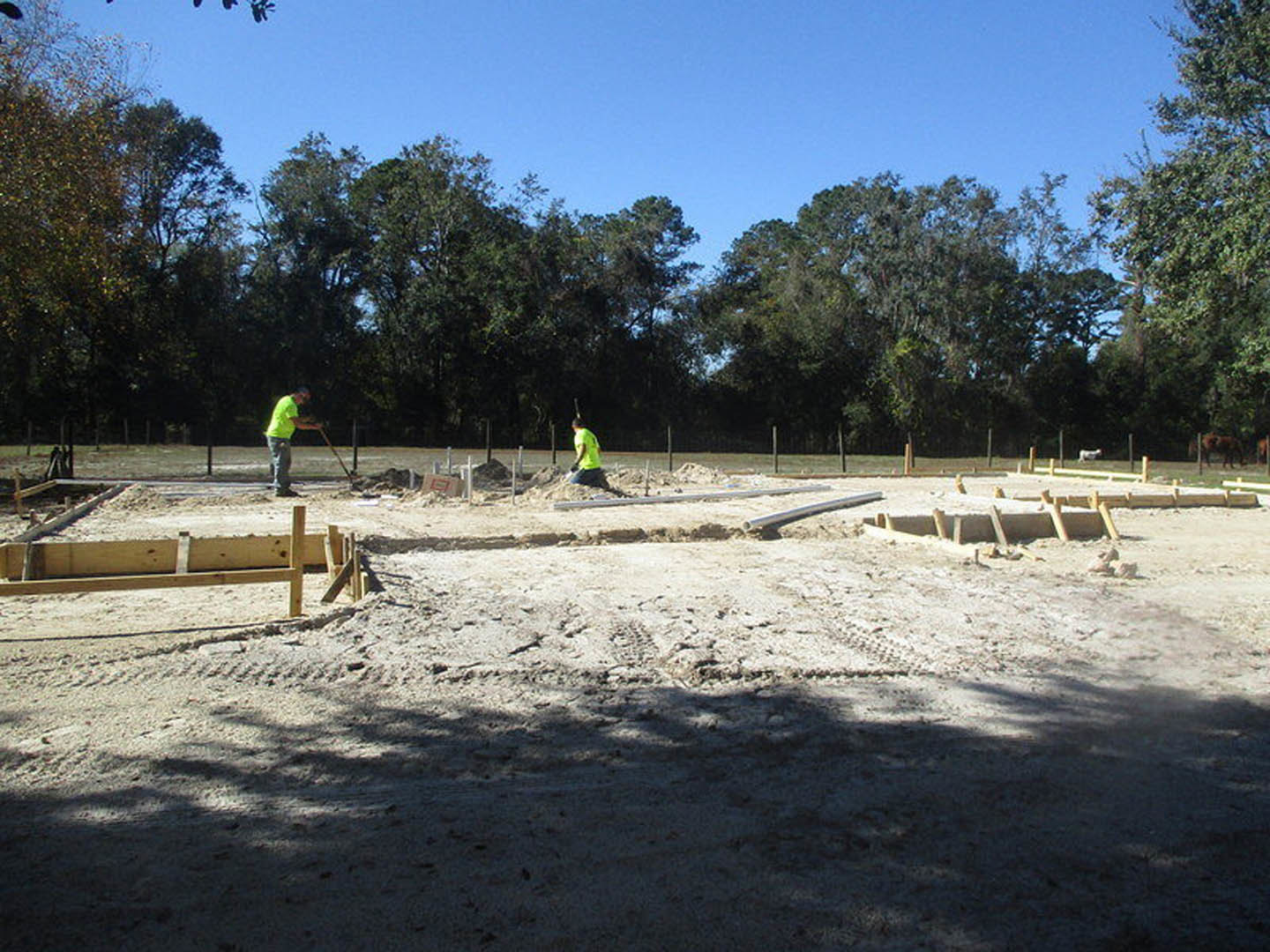 Two men in green and yellow shirts working outdoors at a residential construction site with wooden beams on a dirt surface, trees and sky in the background