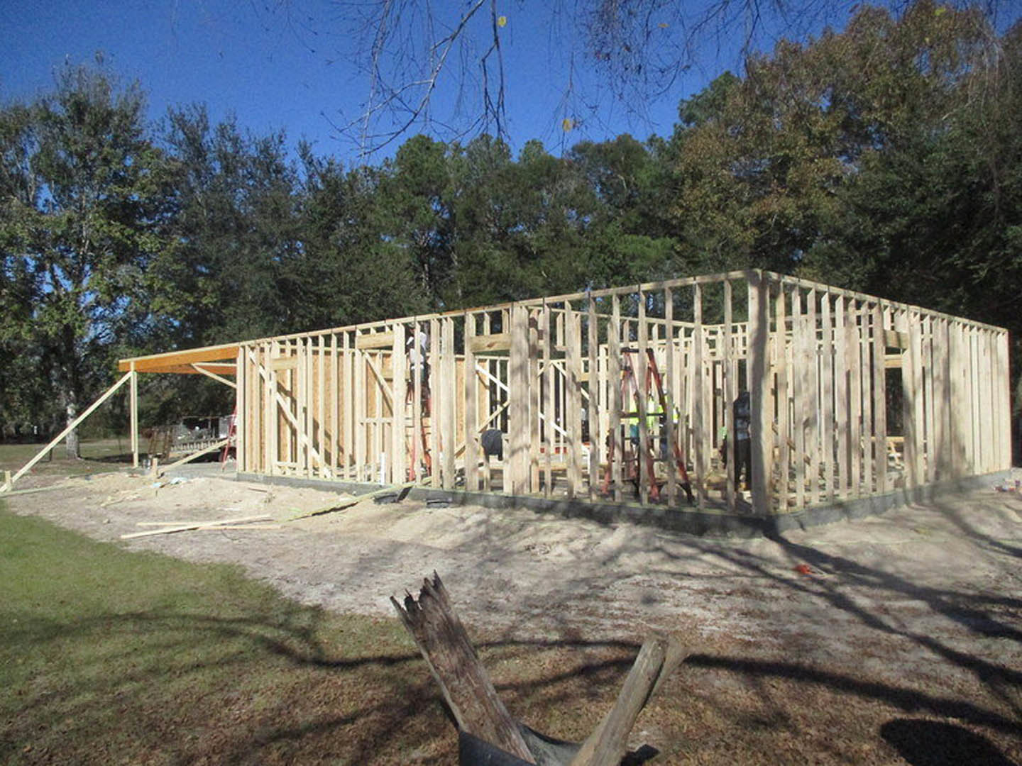 Wood-framed house under construction with exposed beams and plywood, surrounded by dirt and tree stumps, trees and blue sky in background