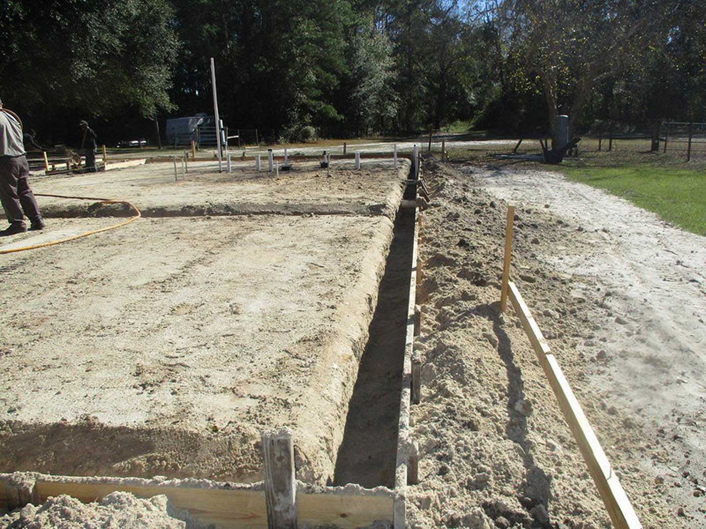Dirt trench dug for home foundation surrounded by soil, grass, and trees; person in brown shorts stands beside excavation on construction site