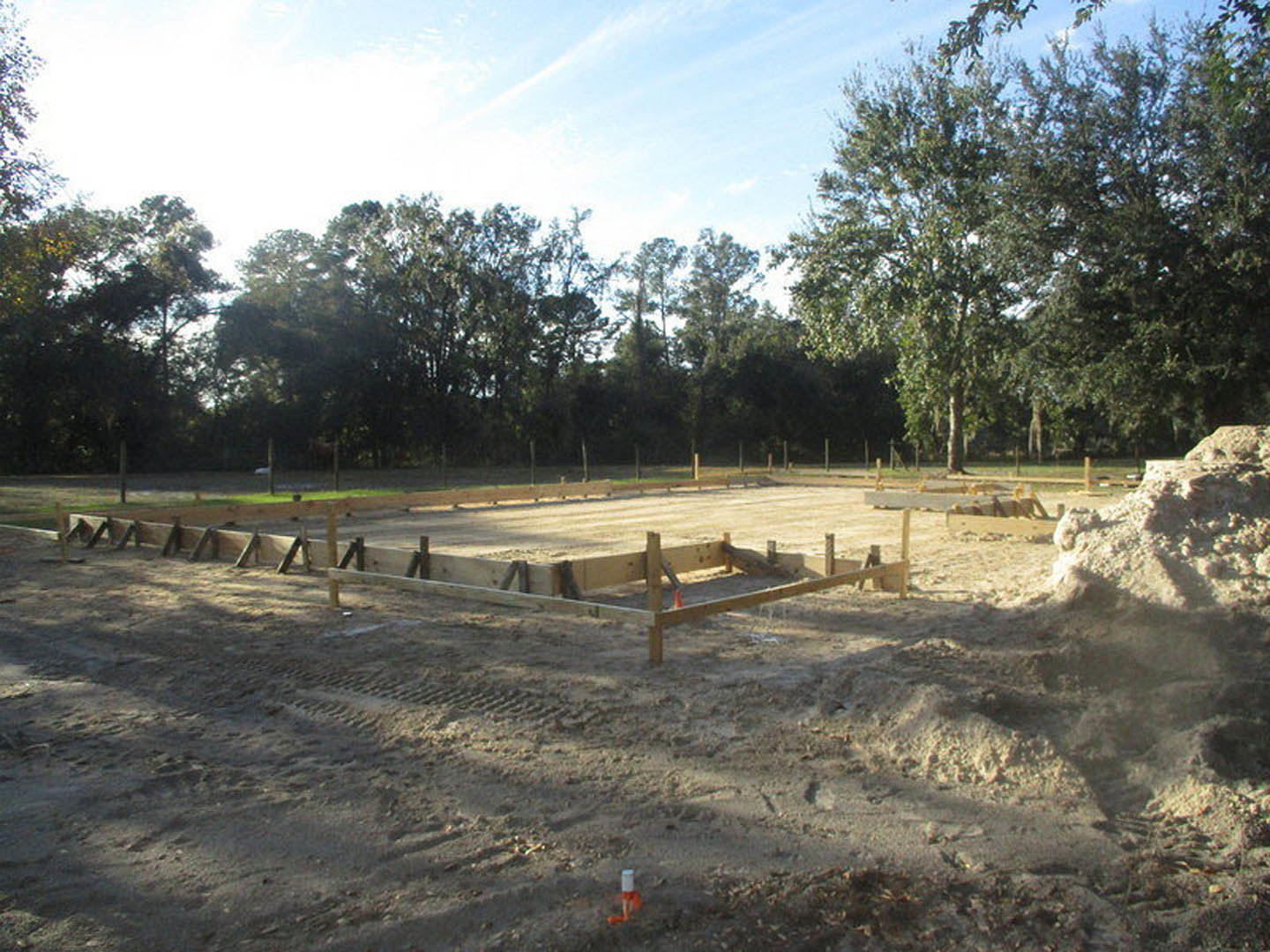 Concrete foundation surrounded by dirt and wooden fencing, pile of soil in foreground, blue sky with scattered clouds above leafy trees in background